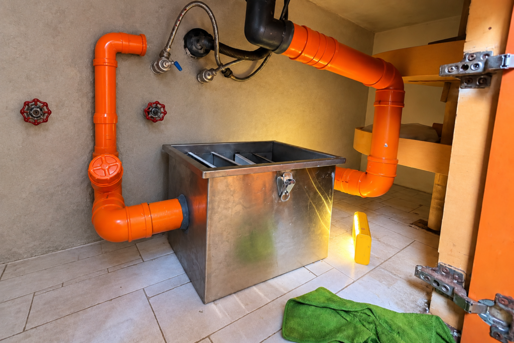 Newly installed stainless steel grease trap connected to orange PVC pipes under a kitchen counter