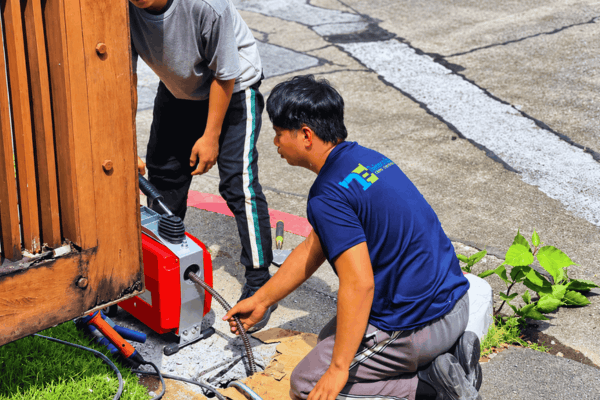 Two Tubero Experts plumbers running a drain cleaning machine near a driveway
