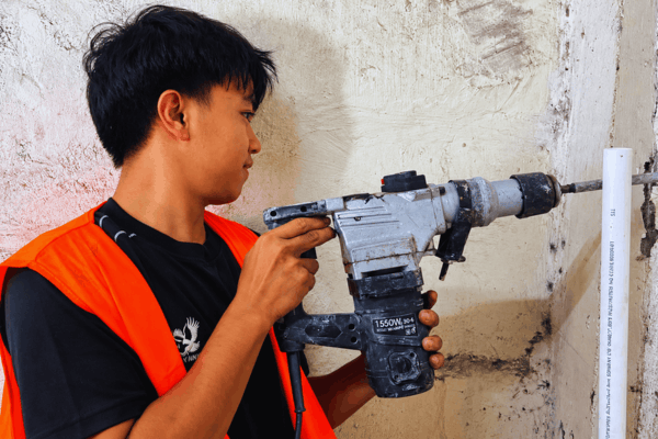 Plumber in an orange safety vest using a rotary hammer to drill into a concrete wall beside a white pipe