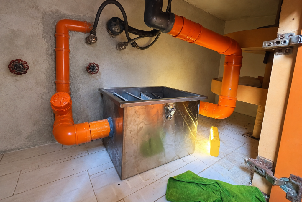 Stainless steel grease trap connected to orange pipes under a kitchen sink