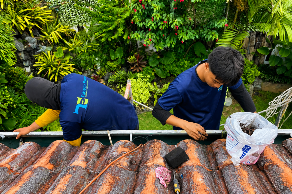 Two Tubero Experts technicians removing debris from a roof gutter on a tiled roof