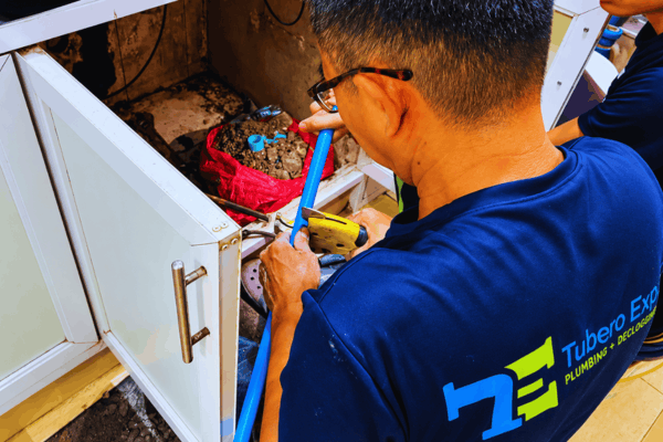 Plumber in glasses cutting a blue water pipe inside an open kitchen cabinet