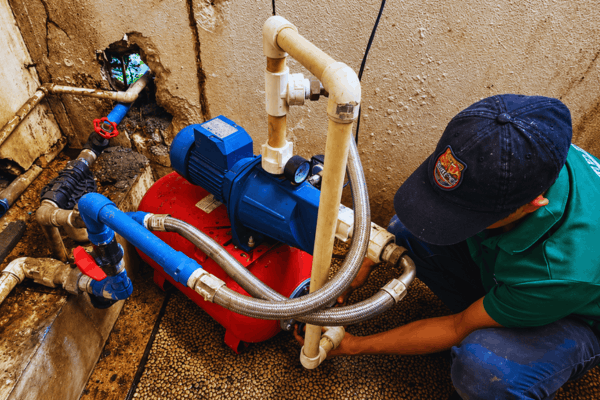 Plumber tightening a fitting on a blue water pump connected to a red pressure tank with braided hoses
